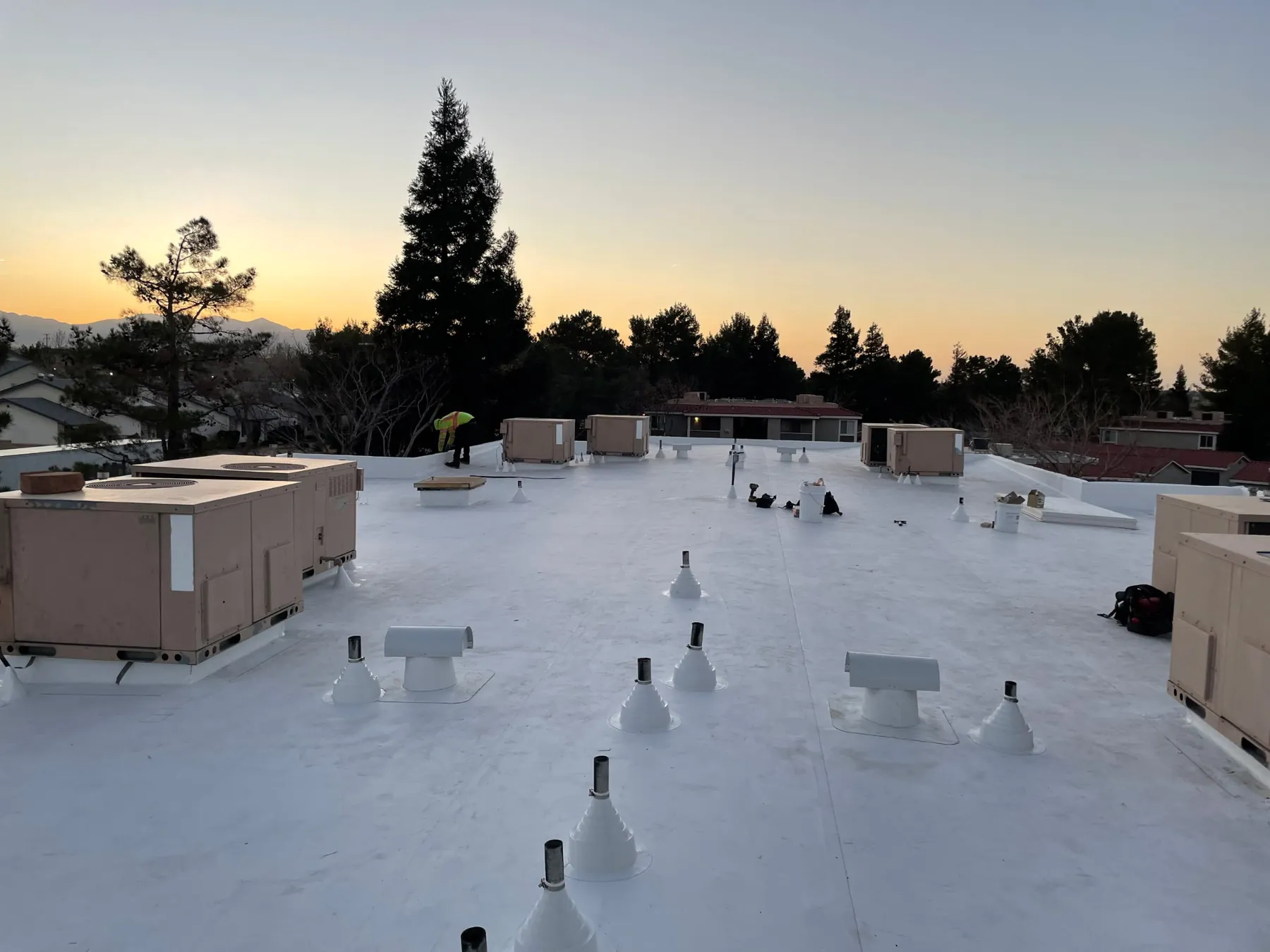 New white flat roof on an apartment building, lined with pipe vents and rooftop units, trees against a glowing sky