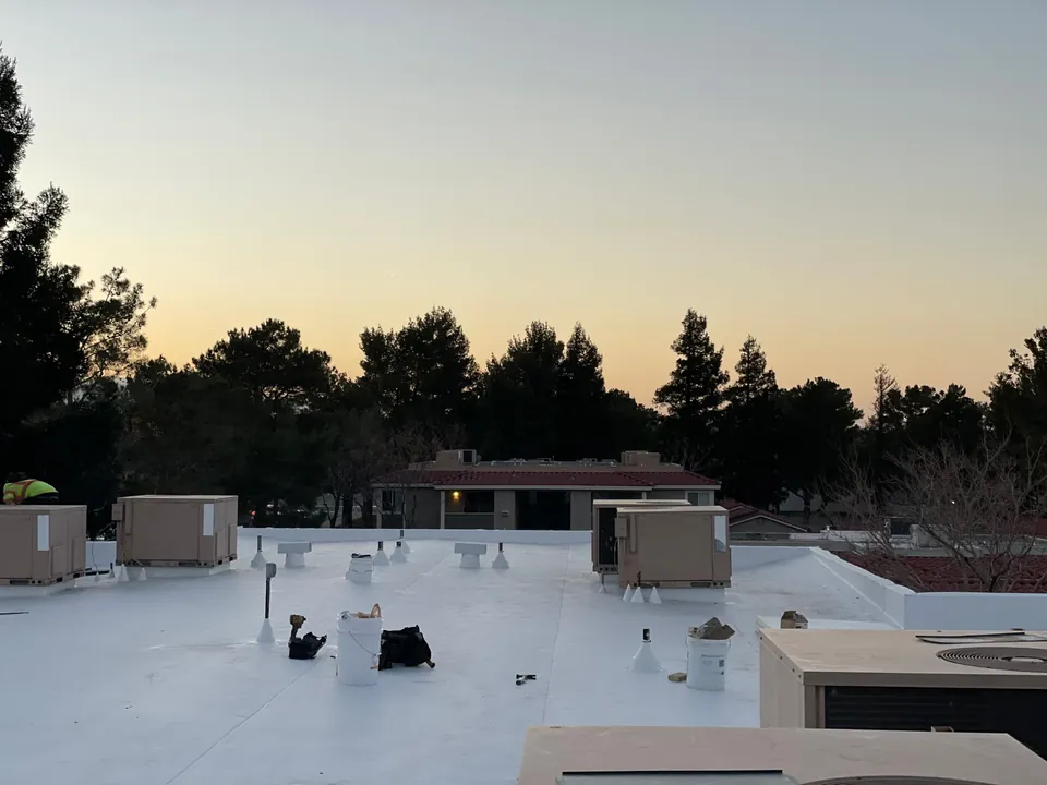 New white flat roof with rooftop units, vents, buckets and tools, trees against a glowing sky