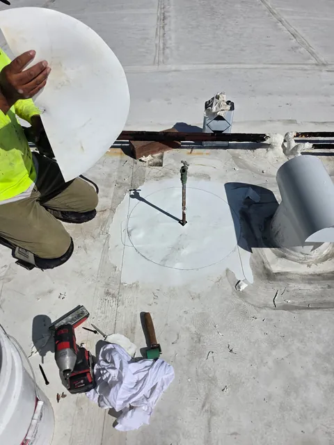 Roofer holding a round membrane patch beside a pipe on a white flat roof; the first patch is already down, with tools nearby